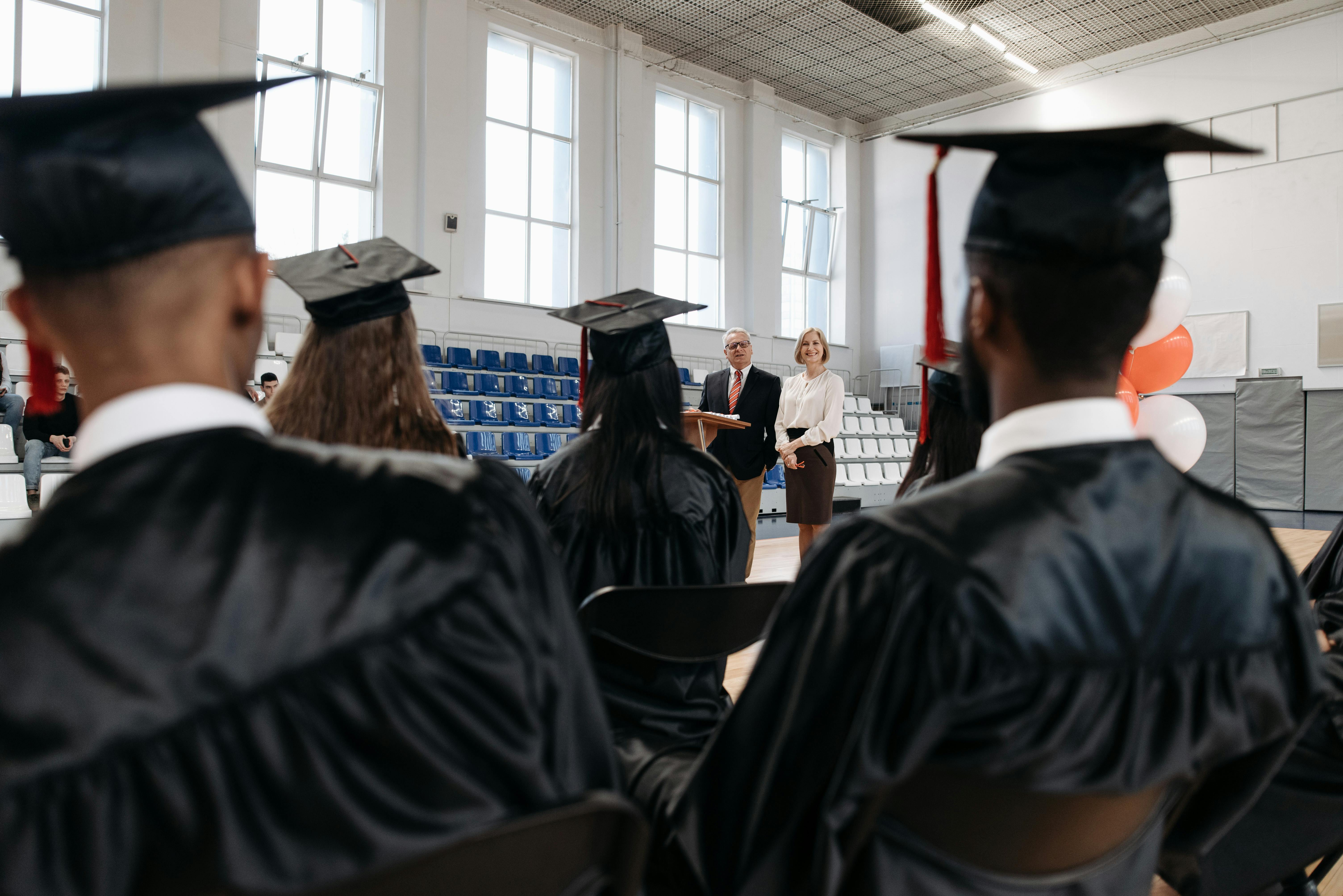 Graduation ceremony with graduates listening to a speaker in an indoor gymnasium setting.